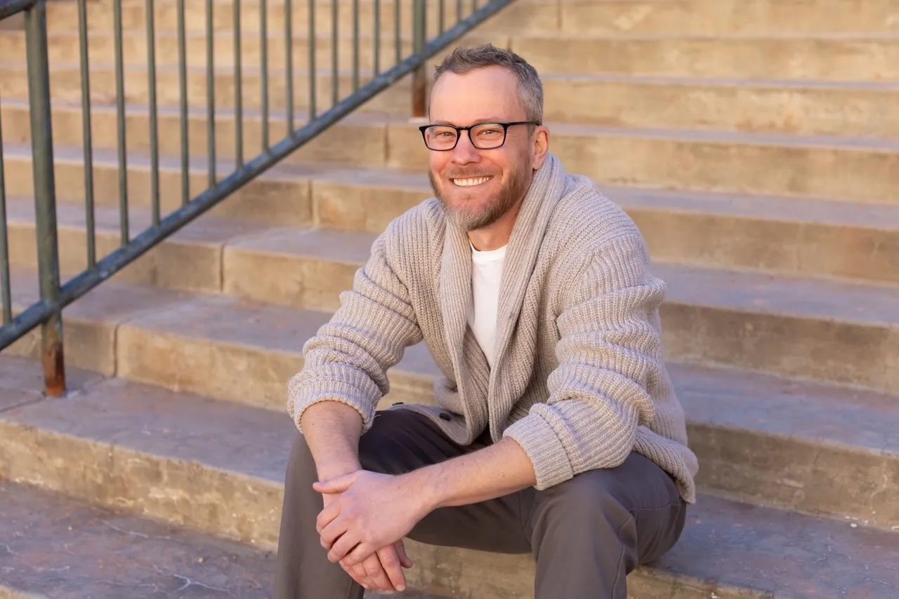 Patrick Maguire, smiling, seated on a flight of stone steps in late afternoon light